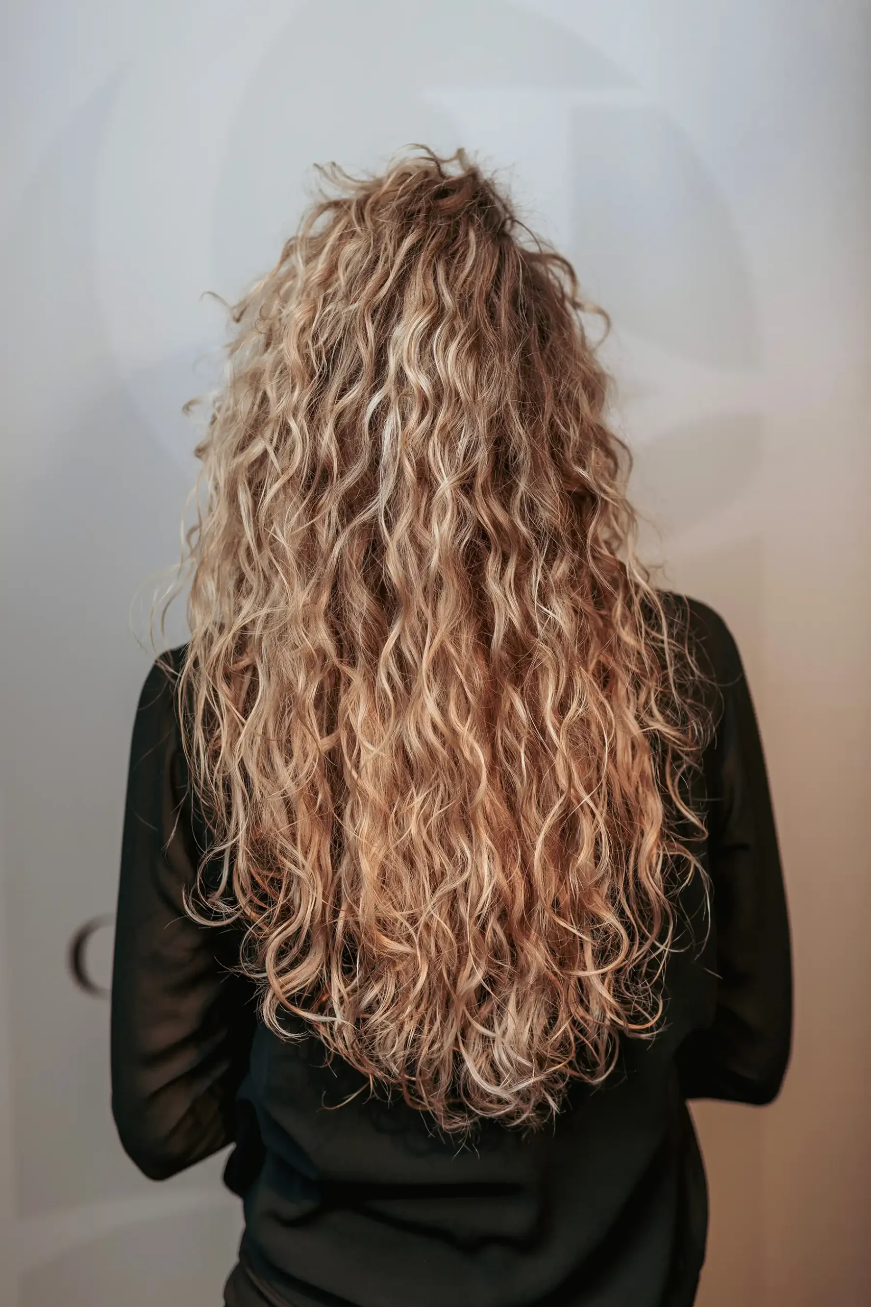Lang, krullend blond haar met een Essential-weaving van Cèlia Blu, van achteren gezien